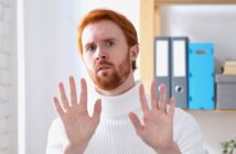 A man with red hair and a beard, wearing a white turtleneck sweater, holds both hands up with palms facing forward, appearing cautious or defensive. Office shelves with binders are visible in the background.