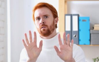 A man with red hair and a beard, wearing a white turtleneck sweater, holds both hands up with palms facing forward, appearing cautious or defensive. Office shelves with binders are visible in the background.