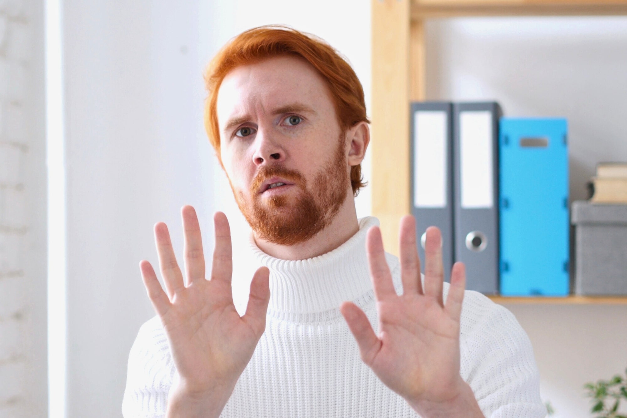 A man with red hair and a beard, wearing a white turtleneck sweater, holds both hands up with palms facing forward, appearing cautious or defensive. Office shelves with binders are visible in the background.