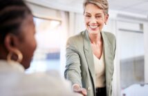 A smiling woman in a light green blazer reaches out to shake hands with another person in an office setting, suggesting a friendly greeting or the start of a business meeting.
