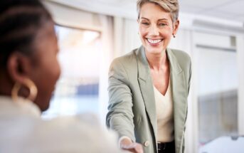 A smiling woman in a light green blazer reaches out to shake hands with another person in an office setting, suggesting a friendly greeting or the start of a business meeting.