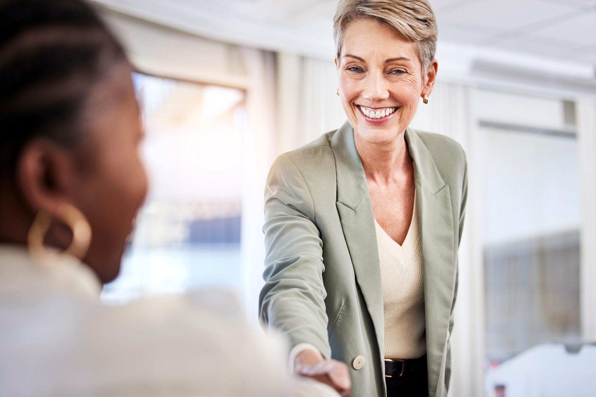 A smiling woman in a light green blazer reaches out to shake hands with another person in an office setting, suggesting a friendly greeting or the start of a business meeting.