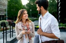 A woman holding a smartphone and a man carrying a bag and laptop smile and talk outdoors in a city park setting, with trees and buildings in the background.