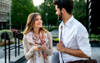 A woman holding a smartphone and a man carrying a bag and laptop smile and talk outdoors in a city park setting, with trees and buildings in the background.