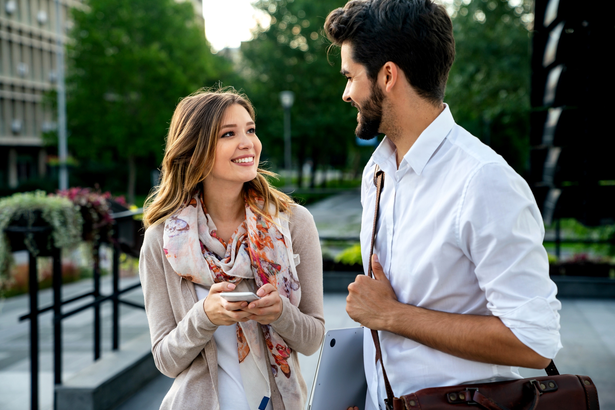 A woman holding a smartphone and a man carrying a bag and laptop smile and talk outdoors in a city park setting, with trees and buildings in the background.