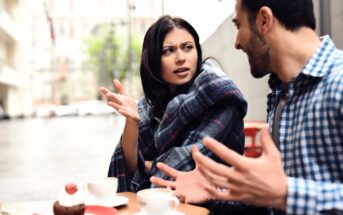 A woman wrapped in a blanket looks frustrated while talking to a man at an outdoor café table with coffee and dessert. Both have expressive hand gestures, suggesting a serious conversation.