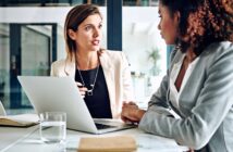 Two women in business attire are sitting at a desk with a laptop and documents, engaged in a serious conversation in a modern office setting.