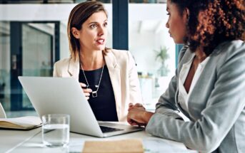 Two women in business attire are sitting at a desk with a laptop and documents, engaged in a serious conversation in a modern office setting.