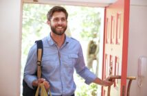 A smiling man with a beard, wearing a blue shirt and carrying a backpack, enters through a red door while holding grocery bags. Sunlight and greenery are visible outside through the open doorway.