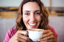 A smiling woman with wavy brown hair holds a cup of coffee with frothy milk, looking directly at the camera. She is wearing a pink top and appears relaxed and happy indoors.