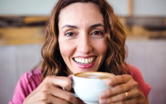 A smiling woman with wavy brown hair holds a cup of coffee with frothy milk, looking directly at the camera. She is wearing a pink top and appears relaxed and happy indoors.