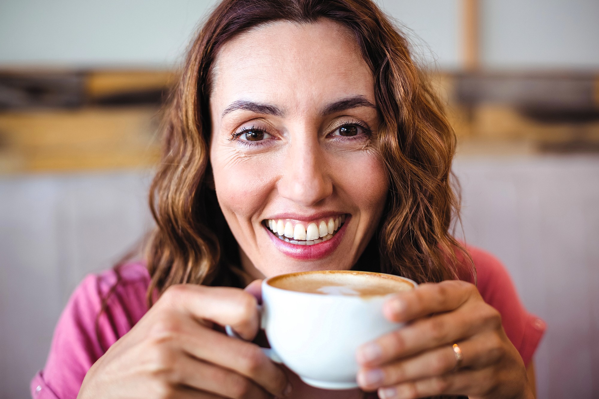 A smiling woman with wavy brown hair holds a cup of coffee with frothy milk, looking directly at the camera. She is wearing a pink top and appears relaxed and happy indoors.