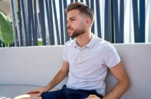 A young man with neatly styled hair and a trimmed beard sits on a light-colored outdoor couch, wearing a pale gray polo shirt and dark shorts, looking thoughtfully to the side. Vertical slats and green plants are in the background.