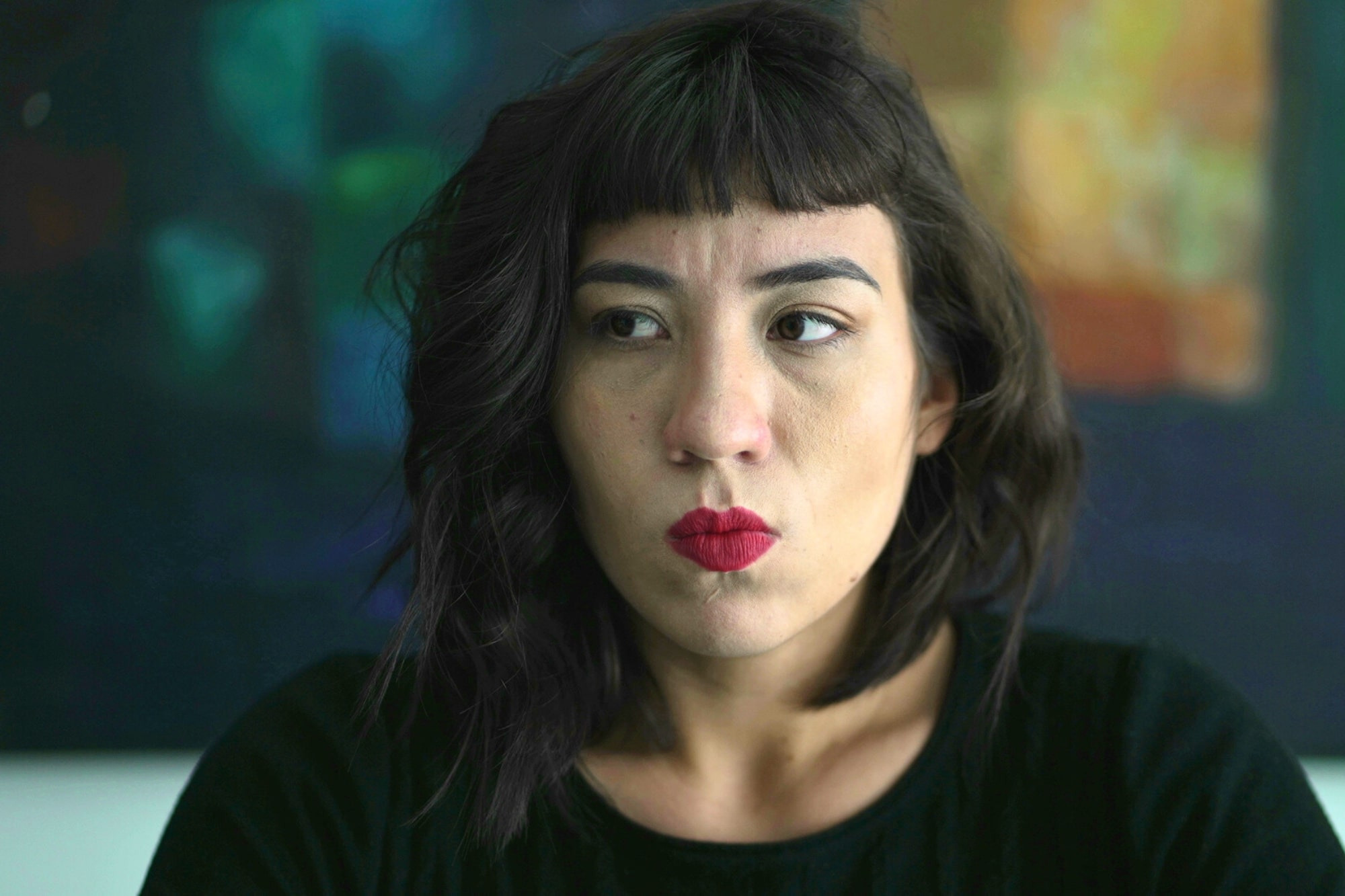 A woman with dark wavy hair and red lipstick looks to the side with a serious expression. She is wearing a black top, and the background is blurred with abstract colors.