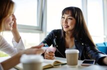 Two women sit at a table in a bright cafe, talking and smiling. They have coffee cups, a notebook, and a smartphone on the table. One woman wears a black jacket; the other gestures with her hand while speaking.