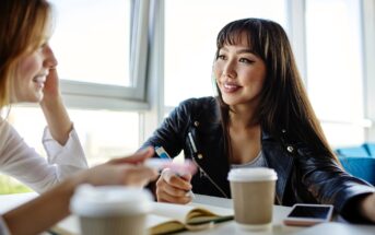 Two women sit at a table in a bright cafe, talking and smiling. They have coffee cups, a notebook, and a smartphone on the table. One woman wears a black jacket; the other gestures with her hand while speaking.
