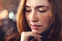 A young woman with long red hair and freckles looks down thoughtfully, resting her chin lightly on her hand. The background is softly blurred with circular light bokeh.