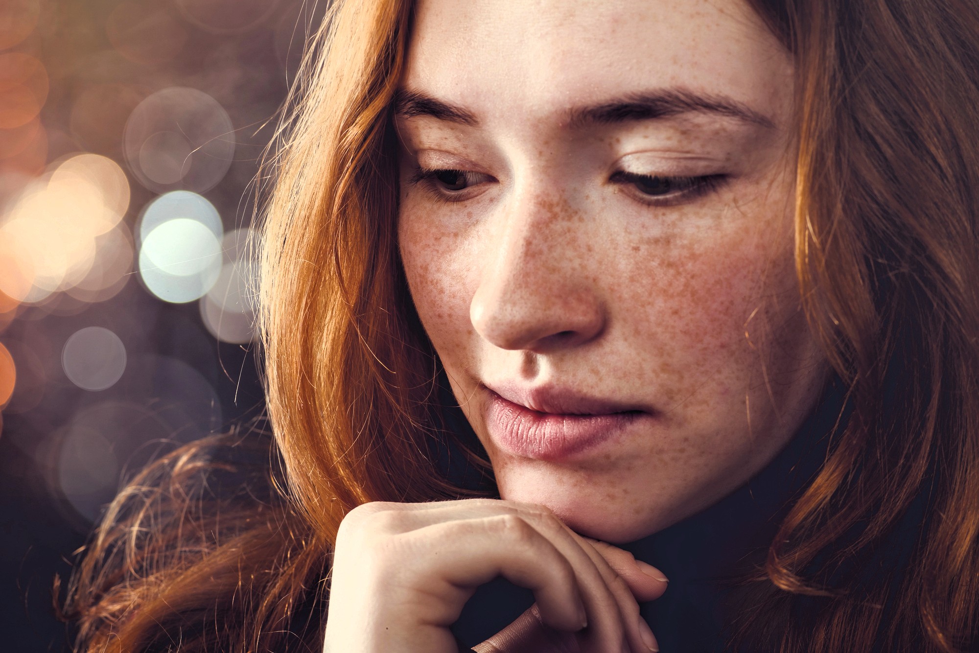 A young woman with long red hair and freckles looks down thoughtfully, resting her chin lightly on her hand. The background is softly blurred with circular light bokeh.