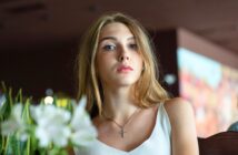 A young woman with long blonde hair wearing a white top and a cross necklace looks at the camera. She is indoors with blurred plants and colorful background elements visible.