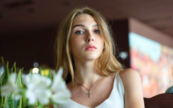A young woman with long blonde hair wearing a white top and a cross necklace looks at the camera. She is indoors with blurred plants and colorful background elements visible.