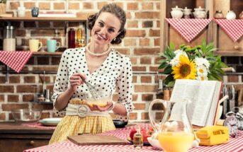 A woman in retro clothing whisks eggs in a kitchen decorated with red checkered cloths and flowers. There's an open cookbook, a glass pitcher, and a vintage phone on the counter. She smiles at the camera.