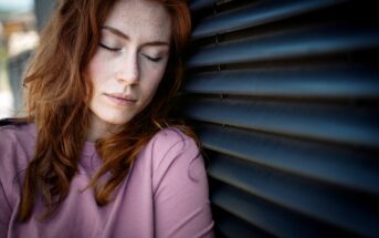 A young woman with long red hair and freckles, wearing a mauve shirt, rests her head and closes her eyes against a dark ribbed wall, looking calm and introspective.