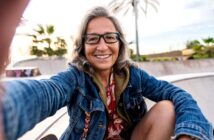 Smiling older woman with long gray hair and glasses takes a selfie at a skatepark, wearing a denim jacket and patterned shirt. Palm trees and a bright sky are visible in the background.