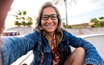 Smiling older woman with long gray hair and glasses takes a selfie at a skatepark, wearing a denim jacket and patterned shirt. Palm trees and a bright sky are visible in the background.
