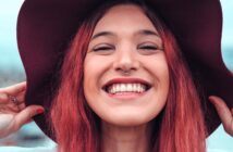 A young person with long red hair smiles widely, showing teeth, while holding the brim of a wide dark hat. They have a nose ring and are outdoors with a blurred background.
