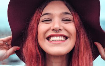 A young person with long red hair smiles widely, showing teeth, while holding the brim of a wide dark hat. They have a nose ring and are outdoors with a blurred background.