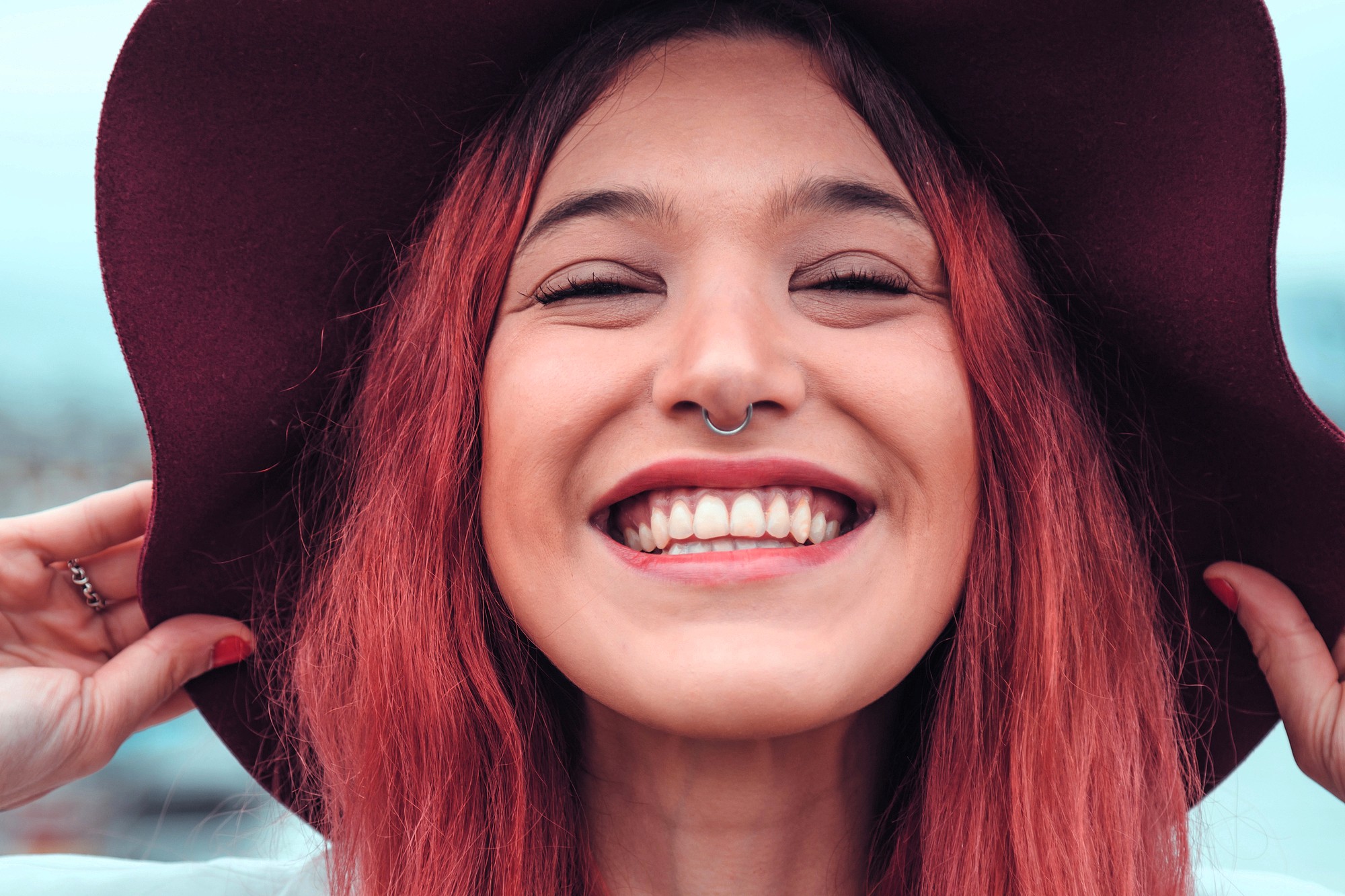 A young person with long red hair smiles widely, showing teeth, while holding the brim of a wide dark hat. They have a nose ring and are outdoors with a blurred background.