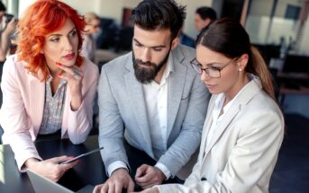 Three business professionals in formal attire are gathered around a laptop, discussing something on the screen. One woman holds a tablet, while the man and another woman focus intently on the laptop.