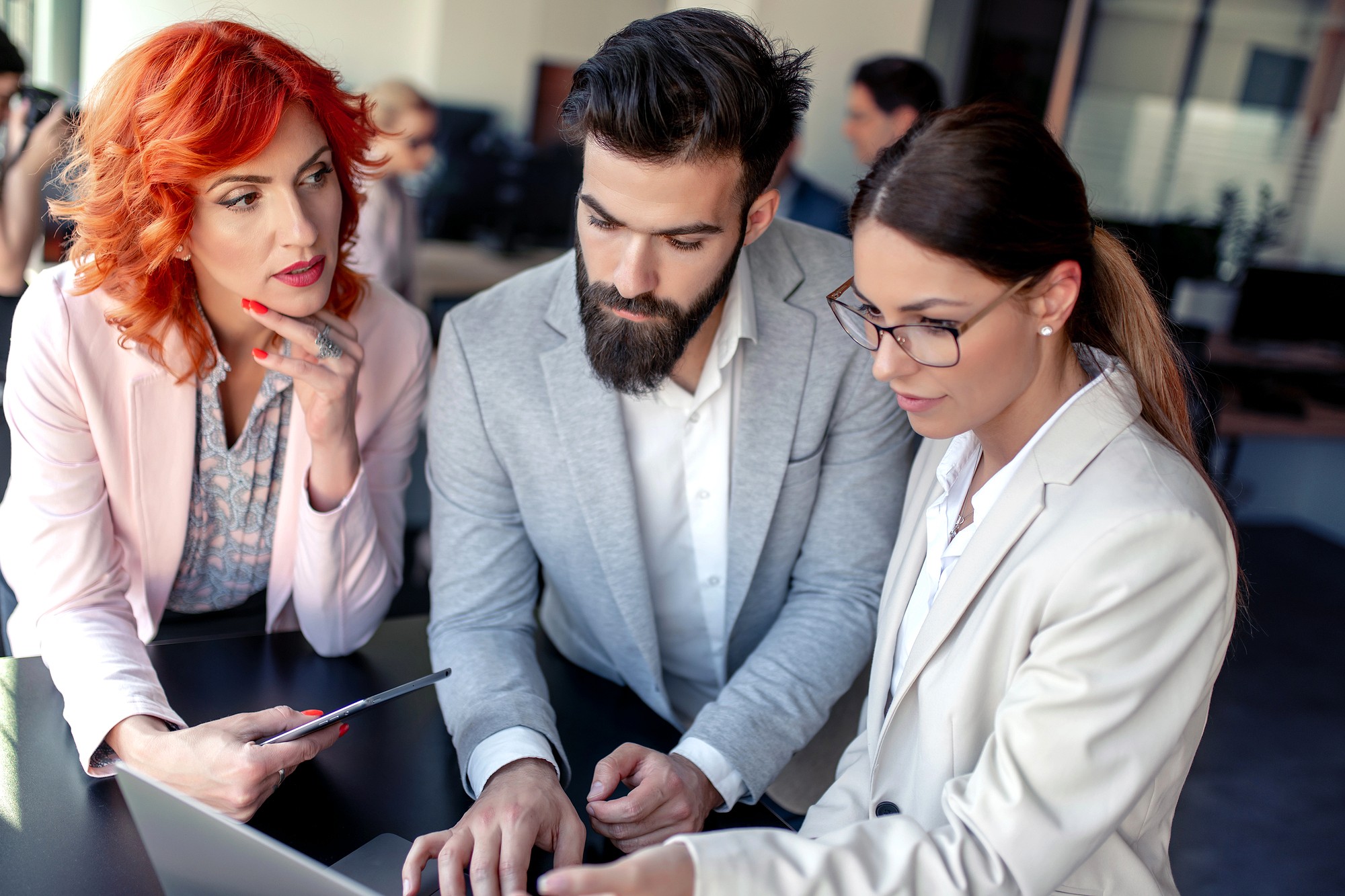 Three business professionals in formal attire are gathered around a laptop, discussing something on the screen. One woman holds a tablet, while the man and another woman focus intently on the laptop.