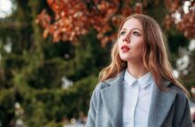 A young woman with long, light brown hair wearing a gray coat and white shirt looks up thoughtfully outdoors. Autumn leaves and green trees are blurred in the background.