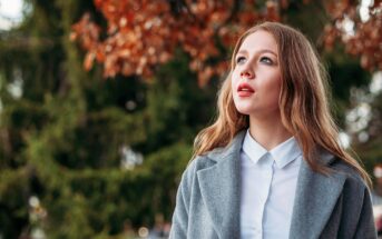 A young woman with long, light brown hair wearing a gray coat and white shirt looks up thoughtfully outdoors. Autumn leaves and green trees are blurred in the background.