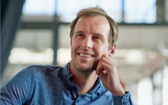 A man with light brown hair wearing a blue button-up shirt is smiling while resting his chin on his hand. The background is blurred with natural light coming through large windows.