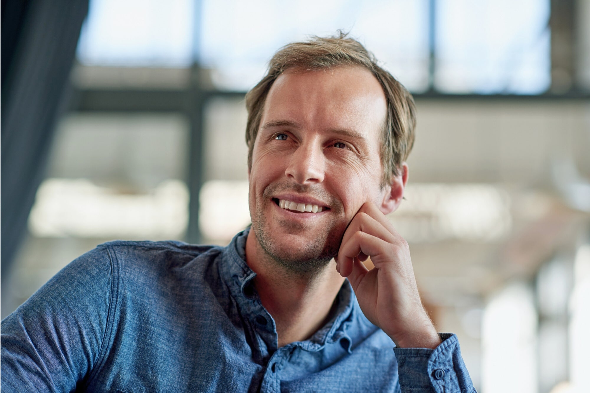 A man with light brown hair wearing a blue button-up shirt is smiling while resting his chin on his hand. The background is blurred with natural light coming through large windows.