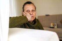 A woman with a serious expression rests her head on her hand while sitting on a couch in a living room, wearing a green sweater. The background shows a beige sofa and soft natural light.