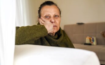 A woman with a serious expression rests her head on her hand while sitting on a couch in a living room, wearing a green sweater. The background shows a beige sofa and soft natural light.
