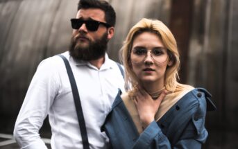 A man with a beard and sunglasses, wearing a white shirt and suspenders, stands next to a woman with blond hair and large glasses in a blue off-shoulder jacket. Both have serious expressions and are outdoors.