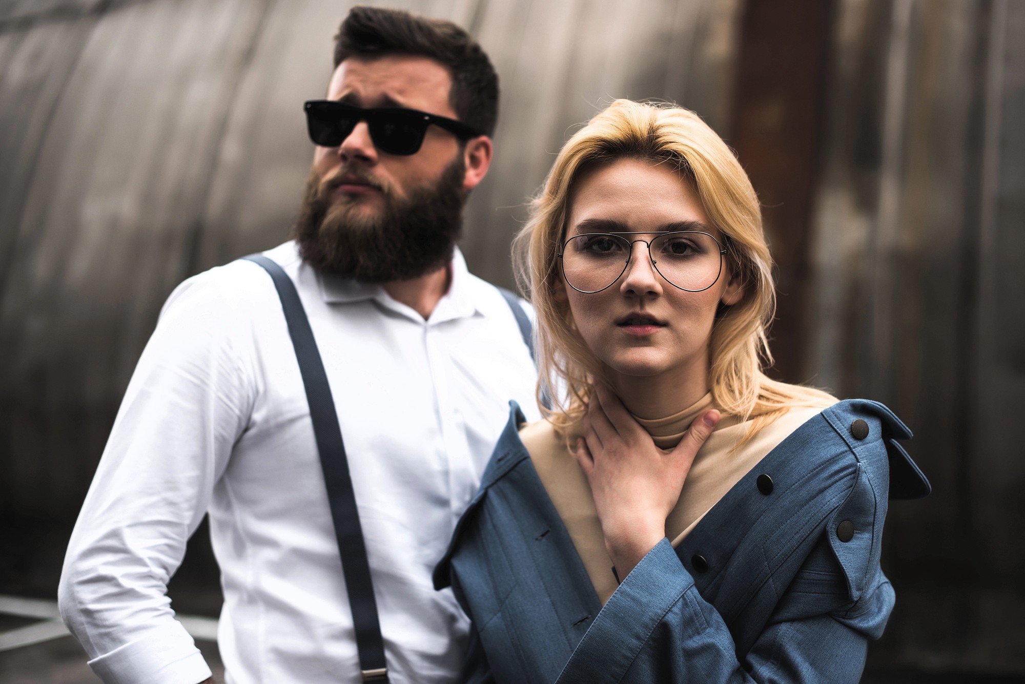 A man with a beard and sunglasses, wearing a white shirt and suspenders, stands next to a woman with blond hair and large glasses in a blue off-shoulder jacket. Both have serious expressions and are outdoors.