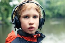 A young child with blonde hair wears large black headphones and a navy blue jacket with an orange hood, standing outdoors near a body of water with blurred green trees in the background.