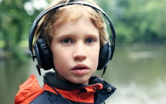 A young child with blonde hair wears large black headphones and a navy blue jacket with an orange hood, standing outdoors near a body of water with blurred green trees in the background.