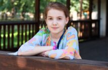 A young girl with light brown hair, wearing a colorful paisley-patterned shirt, leans on a wooden railing and smiles slightly. Trees and a wooden structure appear blurred in the background.