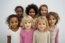 Seven young girls stand close together, smiling at the camera. They have different hair types and colors, and wear light-colored tops, with one in a bright pink shirt. The background is plain and white.