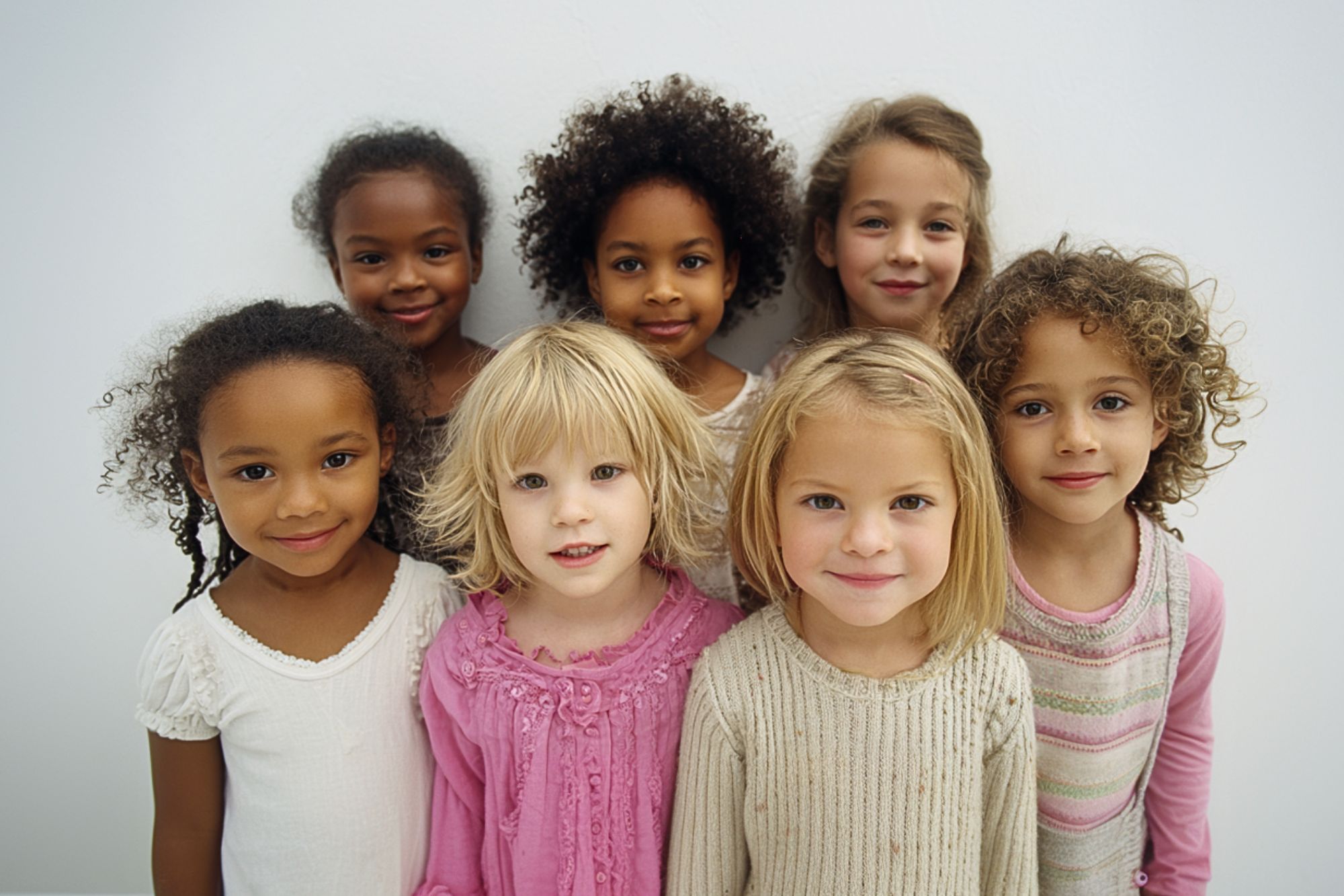 Seven young girls stand close together, smiling at the camera. They have different hair types and colors, and wear light-colored tops, with one in a bright pink shirt. The background is plain and white.