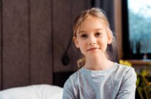 A young girl with blonde hair, wearing a light gray long-sleeve shirt, sits indoors with a calm expression. The background features a bed, a dark wall, and a window with daylight coming through.