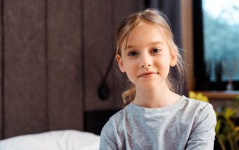 A young girl with blonde hair, wearing a light gray long-sleeve shirt, sits indoors with a calm expression. The background features a bed, a dark wall, and a window with daylight coming through.