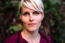 A woman with short blonde hair and blue eyes looks directly at the camera, wearing a burgundy shirt, with a blurred outdoor background of greenery.