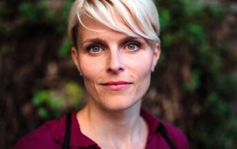 A woman with short blonde hair and blue eyes looks directly at the camera, wearing a burgundy shirt, with a blurred outdoor background of greenery.